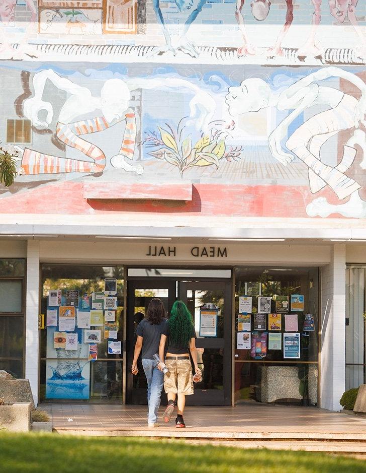 two students walk towards the entrance of mead hall with a bright mural above them