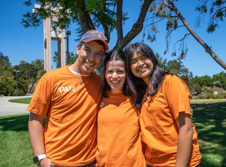 three student admission staff wearing orange tshirts pose on the mounds with the clock tower behind them